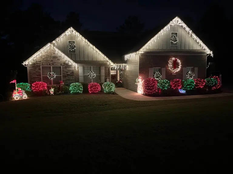 Home at night with white icicle-style lights along the rooflines, illuminated wreaths, and red and green lights on front shrubs.